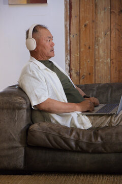 Senior Asian man sitting on dark brown leather couch wearing headphones and typing on laptop