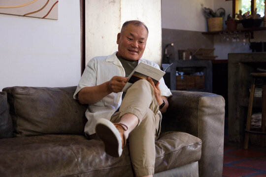 Senior Asian man sitting on leather sofa holding paperback and wearing short-sleeve shirt at home