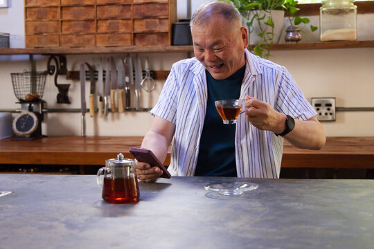 Senior male standing behind metal counter in kitchen, holding glass teacup and checking smartphone