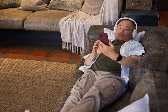 Senior Asian man reclining on right leather sofa, holding red phone, wearing headphones, copy space