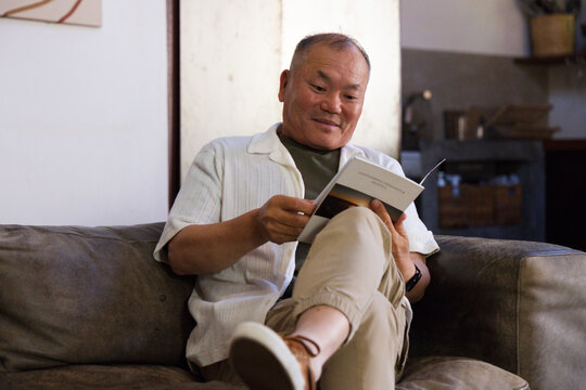 Senior Asian man sitting on couch reading paperback book in living room, wearing wristwatch