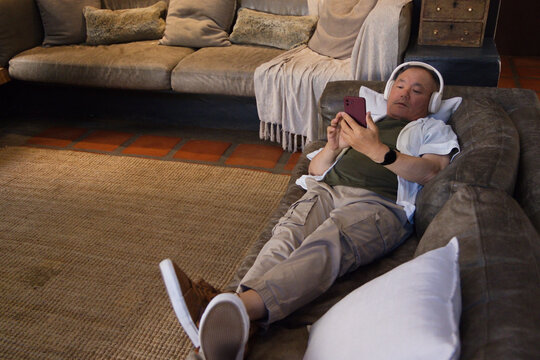 Senior Asian man resting on chaise at home, wearing headphones and holding smartphone, copy space