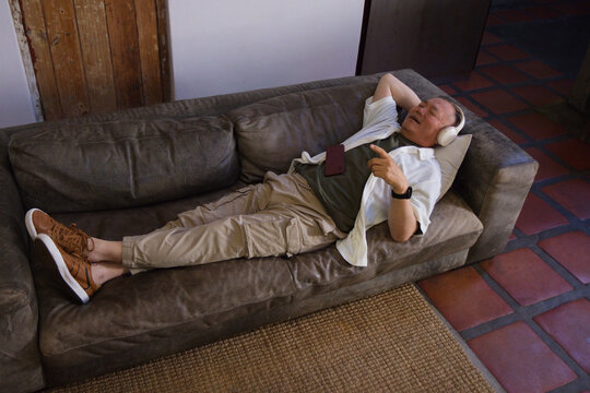 Senior Asian man in casual shirt reclining on sofa at home listening with headset pointing phone