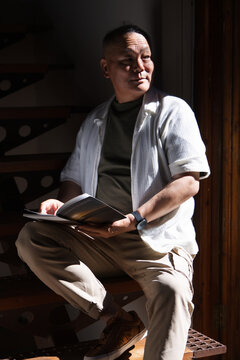 Senior Asian man sitting on metal stair bench reading open book, wearing khaki pants and wristwatch