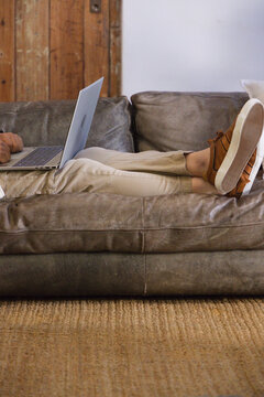 Senior man reclining on grey leather couch using laptop in living room with brown sneakers