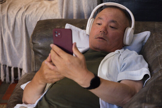 Senior Asian man reclining on leather couch at home, wearing headphones, holding maroon smartphone