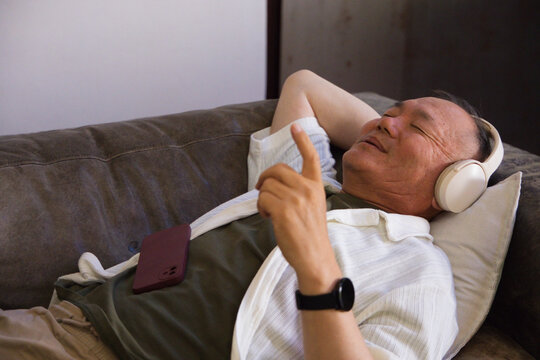 Senior Asian man reclining on dark-gray couch at home wearing cream headphones and tapping phone