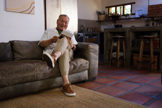 Senior Asian man sitting reading paperback on leather sofa in living room near kitchen counter