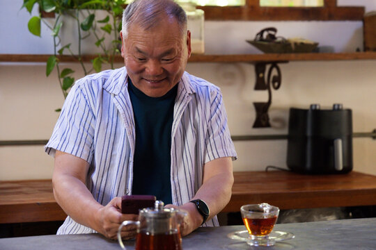 Senior Asian man wearing striped shirt and watch sitting at kitchen bar holding smartphone, teacup