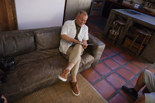 Senior Asian man sitting on brown suede couch in living room opening into kitchen, reading magazine