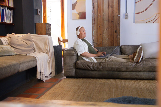 Senior Asian man reclining on grey leather sofa at home, wearing headphones and using laptop