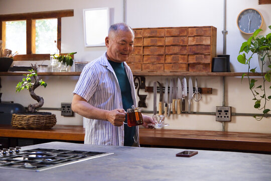 Senior Asian man pouring tea at kitchen counter from glass teapot into clear cup