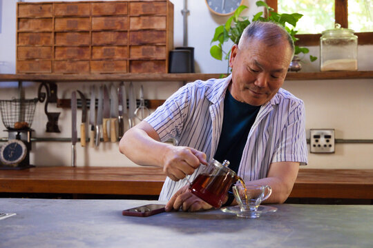 Senior Asian man pouring amber tea from teapot into double-wall cup beside smartphone in kitchen