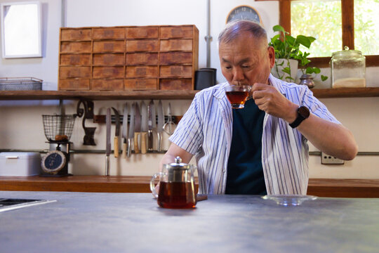 Senior Asian man sipping tea from glass teacup at home kitchen counter with glass teapot