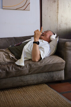 Senior Asian man reclining on brown sofa in living room, wearing headphones, holding red smartphone