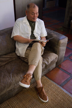 Senior Asian man sitting on worn brown suede sofa at home, reading paperback book