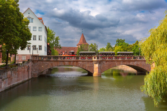 Stone bridge over the river in Nuremberg, Germany, with a tourist train, historic architecture and green trees.