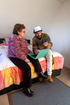 An adult man helps his elderly mother complete online tasks using his cell phone and a laptop