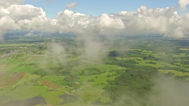 Flying through wispy clouds reveals expansive green landscape under bright blue sky with fluffy white clouds.