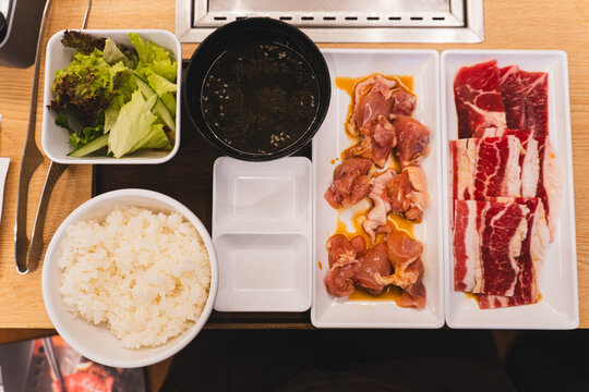 Top down view of a Japanese yakiniku BBQ set meal featuring raw beef slices, marinated chicken, white rice, soup, and salad on a wooden restaurant table