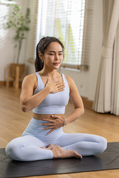 Asian woman practicing yoga meditation indoors with hands on heart and abdomen, calm focus and mindful breathing in bright home studio