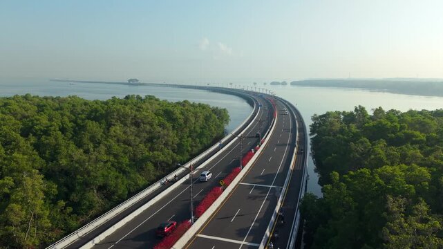 Aerial view of the Bali Mandara Toll Road stretching over the water, surrounded by lush mangrove forests under a clear sky in Kecamatan Kuta Selatan, Bali, Indonesia.