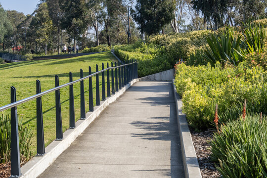 A concrete accessible pedestrian ramp with modern metal handrails curving through landscaped garden beds and grassy parklands in Werribee, Melbourne, Australia.
