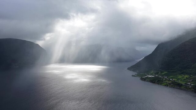 Aerial view of dramatic sunbeams breaking through storm clouds over the dark waters of a fjord with steep mountains and a small village in Valldal, More og Romsdal, Norway.
