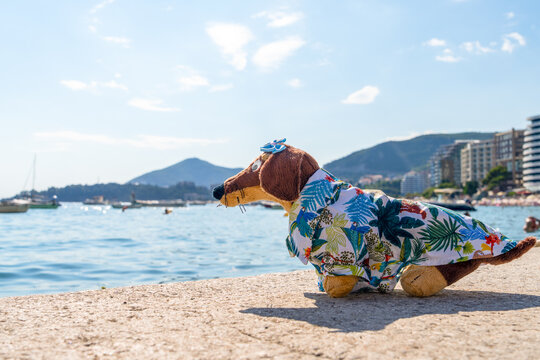 A brown plush dachshund dog toy wearing a tiny blue bow and a tropical shirt sits on a stone ledge above a calm bay, a concept of relaxed seaside daydreaming.