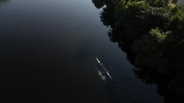 Aerial view of a rowing boat gliding across the calm dark waters of the River Lune, surrounded by lush green trees along the riverbank in Lancaster, England, United Kingdom.