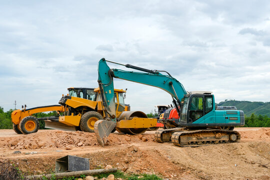 Large yellow and teal excavators and road rollers rest on a construction site during a cloudy afternoon while preparing the ground for building work