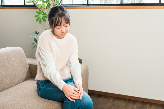 A young girl, either elementary or middle school age, is sitting on the sofa at home, clutching her knee in pain.