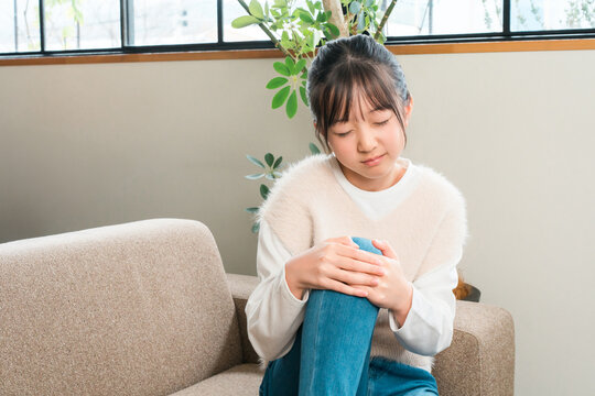 A young girl, either elementary or middle school age, is sitting on the sofa at home, clutching her knee in pain.