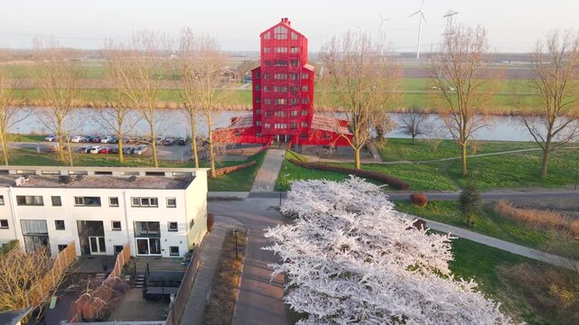 Aerial view of the distinctive red building in the Regenboogbuurt district surrounded by blooming cherry trees, a canal, and modern residential architecture in Almere, Flevoland, Netherlands.