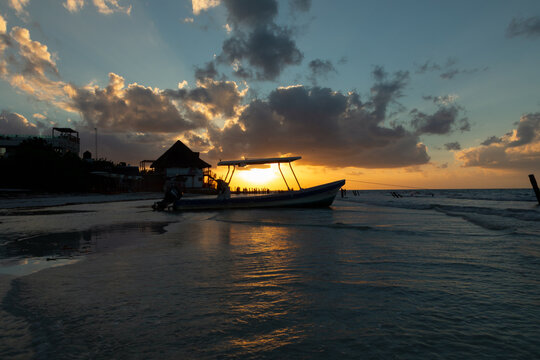 Small fishing boat silhouette on calm beach shallow water under sun breaking through dark clouds.