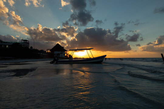 Small fishing boat silhouette on calm beach shallow water under sun breaking through dark clouds.