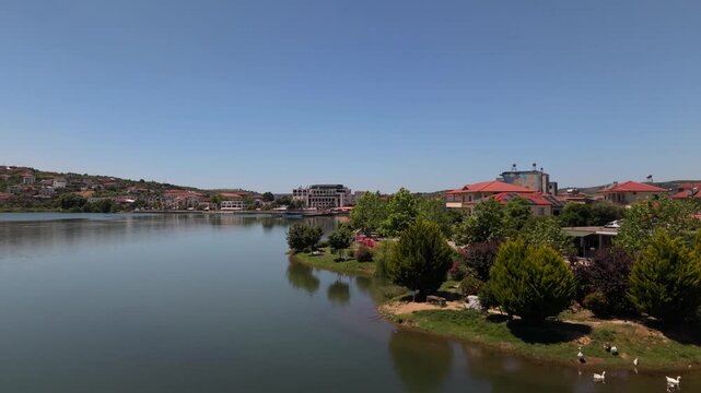 Aerial view of the calm waters of Belsh Lake surrounded by lush trees, red-roofed buildings, and geese on the shore in Belsh, Elbasan County, Albania.