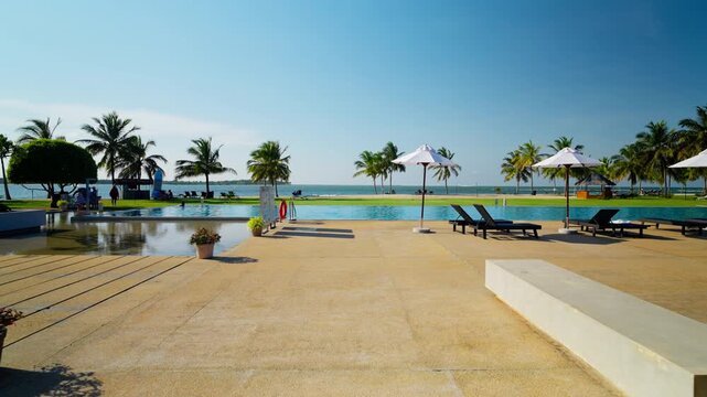 Sun loungers and parasols line an empty resort swimming pool fringed by coconut palms beside a calm turquoise bay in Sri Lanka. The pool looks straight out over the calm bay.