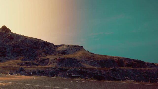 A person walks along a road near mountains under a bright sky. The sun adds light to the scene. The ground is flat with some rocks and grass along the edges. It is a clear day.