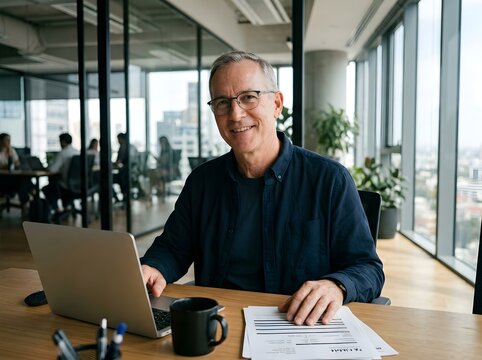 Professional middle-aged man smiling while reviewing documents at a modern glass-walled office desk with laptop and coffee mug