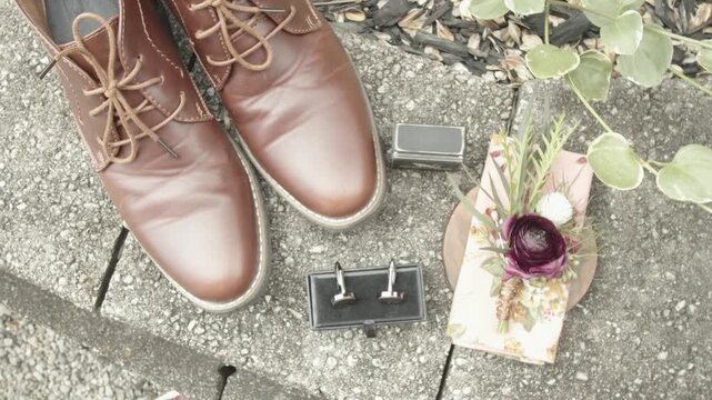 Groom wedding accessories flat lay featuring leather shoes cufflinks and floral boutonniere on stone ground.