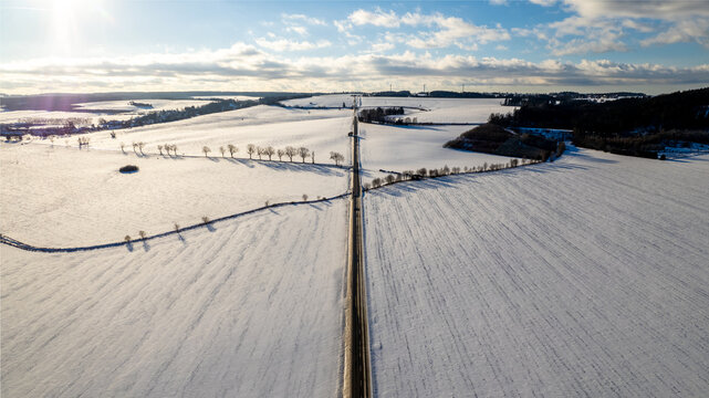 Aerial view of a straight road cutting through vast snow-covered fields under a bright winter sky with distant trees and soft sunlight, Czech Republic.
