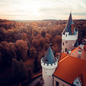 Aerial view of a historic castle with conical towers and orange tiled roofs surrounded by dense autumn forest under a soft sunset sky in CZ.