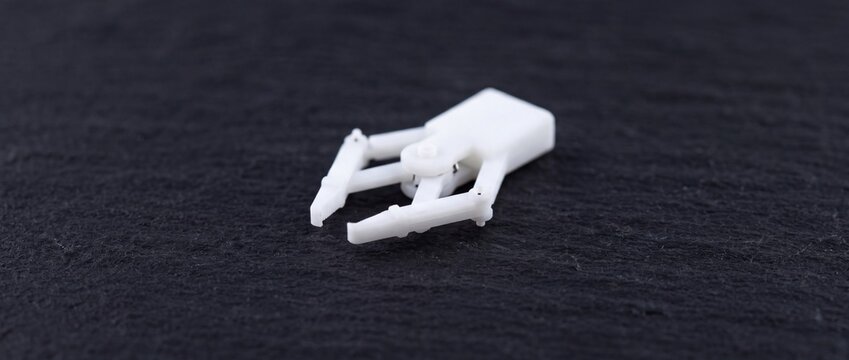 Small white plastic robotic gripper tool with articulated mechanical pincers for precise grasping in automated engineering systems set on dark slate stone surface.