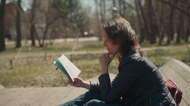 Close up of woman reading book on wooden bench in park with trees background. Book held open fingers resting chin as sunlight filters through branches. Outdoor setting with soft natural