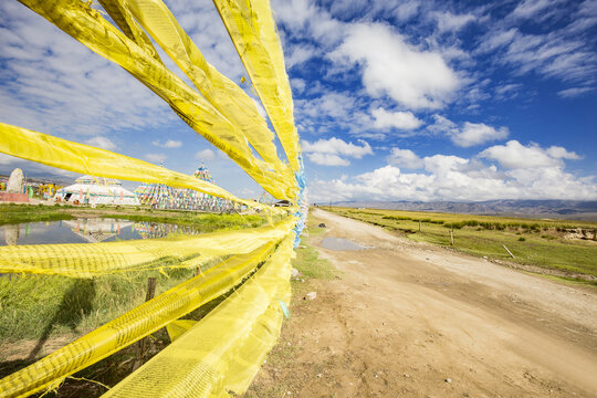 Bright yellow prayer flags stretch along a dirt road in an open grassland, under a blue sky with white clouds.