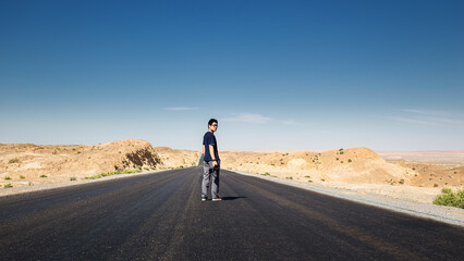 A man stands on a paved road in an arid desert landscape under a clear blue sky, surrounded by rocky terrain. © tai