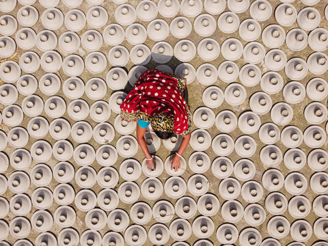 Brahmanbaria, Bangladesh - 03 November 2021: Aerial view of a woman in a red patterned sari arranging numerous unbaked clay pots on the ground to dry in the sun.