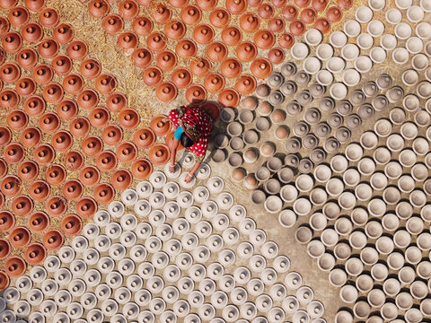 Brahmanbaria, Bangladesh - 03 November 2021: Aerial view of a woman in a red patterned sari arranging rows of terracotta pottery and clay bowls drying on the ground.