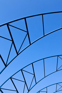 A close-up architectural detail of a modern metal roof truss structure with curved beams and geometric triangular patterns against a clear blue daytime sky.